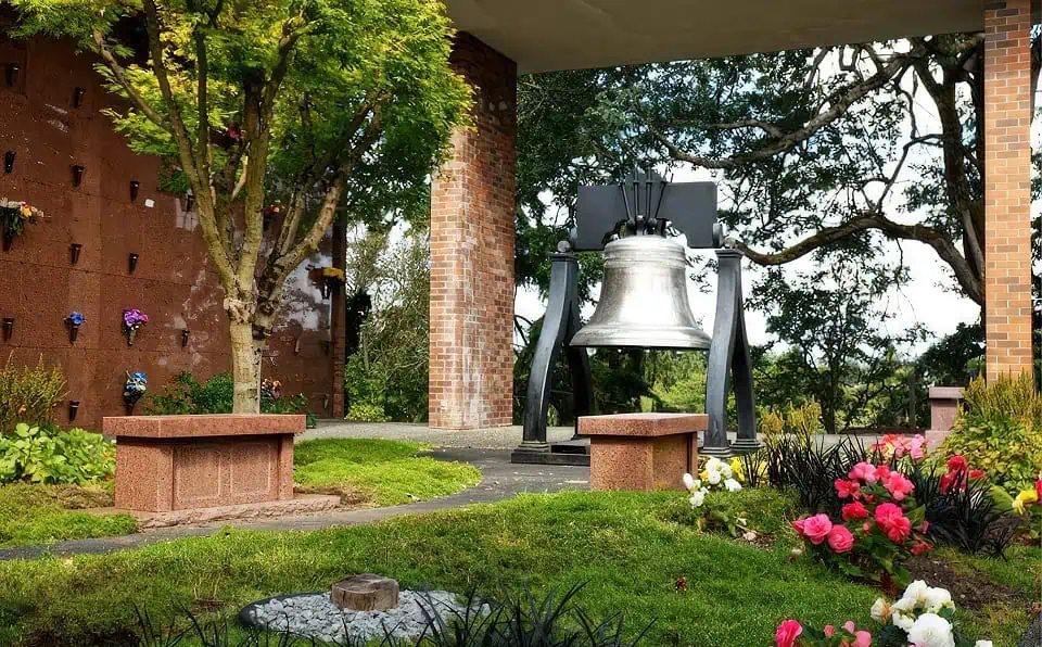 Large bell with black frame in a garden setting with trees and flowers.