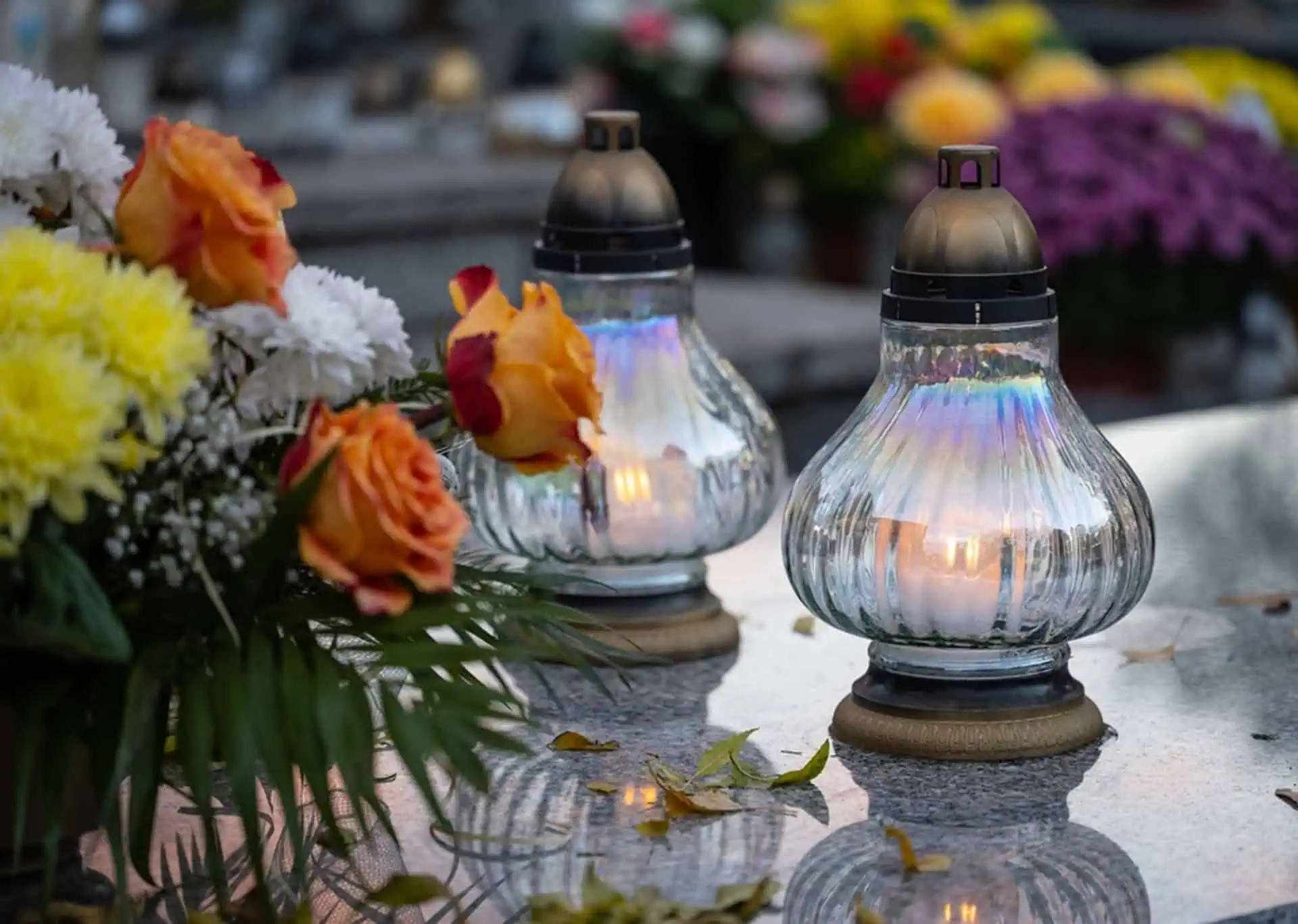 Two glass lanterns with candles on a memorial table surrounded by flowers.