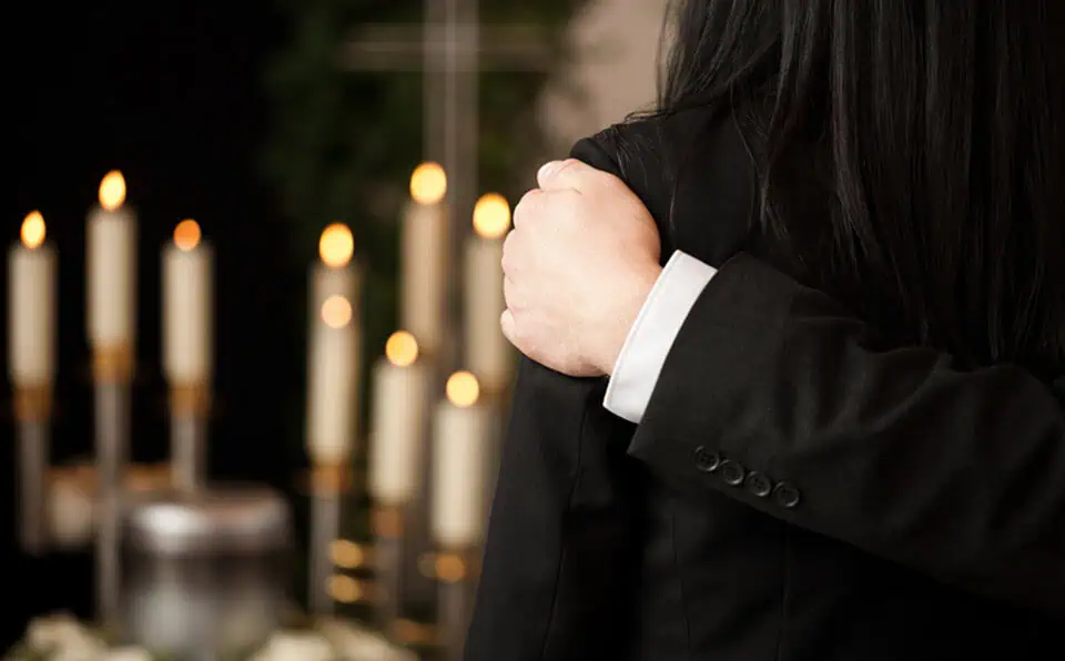 Person in black suit embracing another at a memorial with lit candles in background.