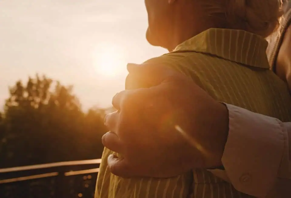 Person being comforted outdoors during sunset, with a caring hand on their shoulder.