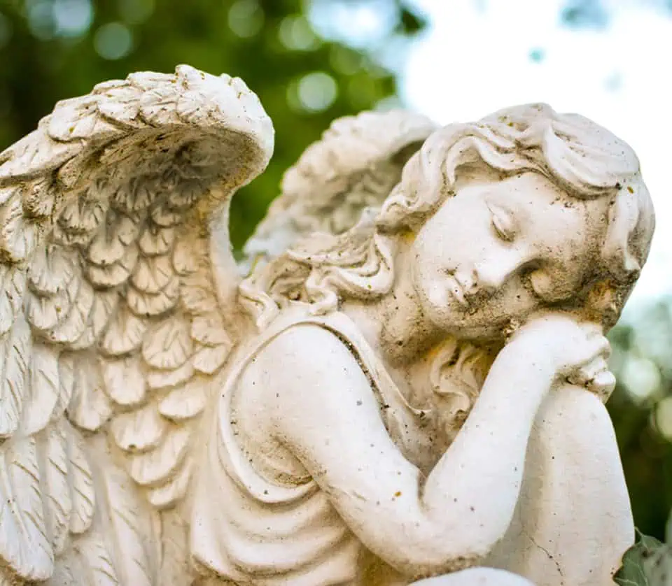 Stone angel statue with closed eyes and folded hands in a garden setting.