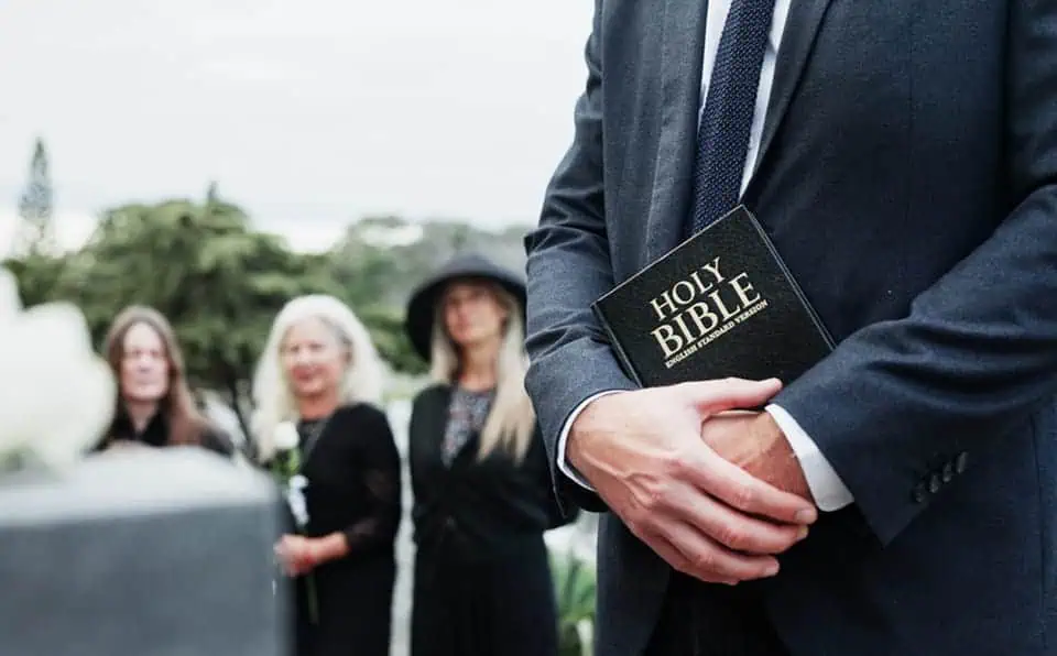 Man in suit holding Holy Bible during outdoor funeral service.