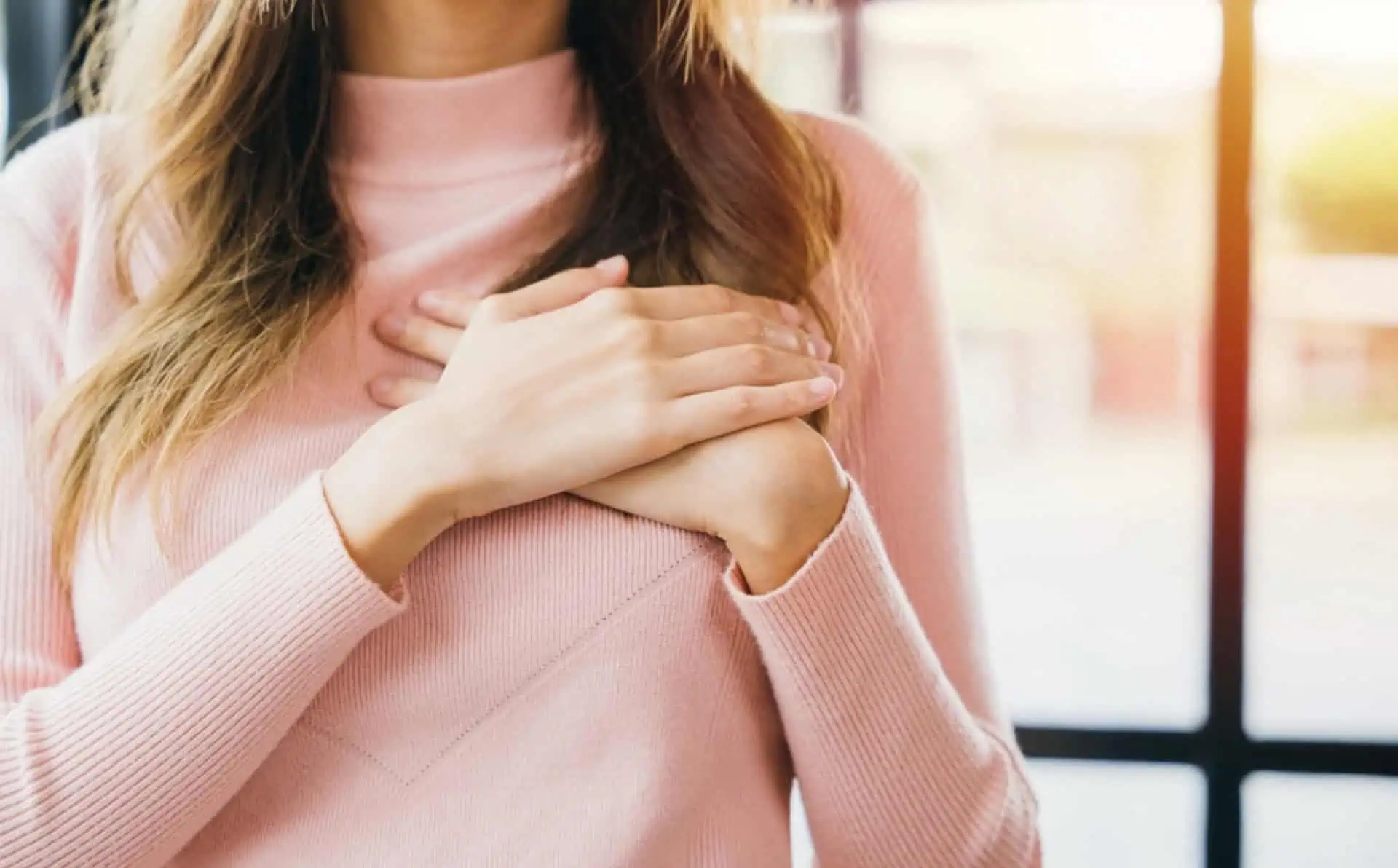 Close-up of woman with hand on chest, wearing pink sweater, indoors.