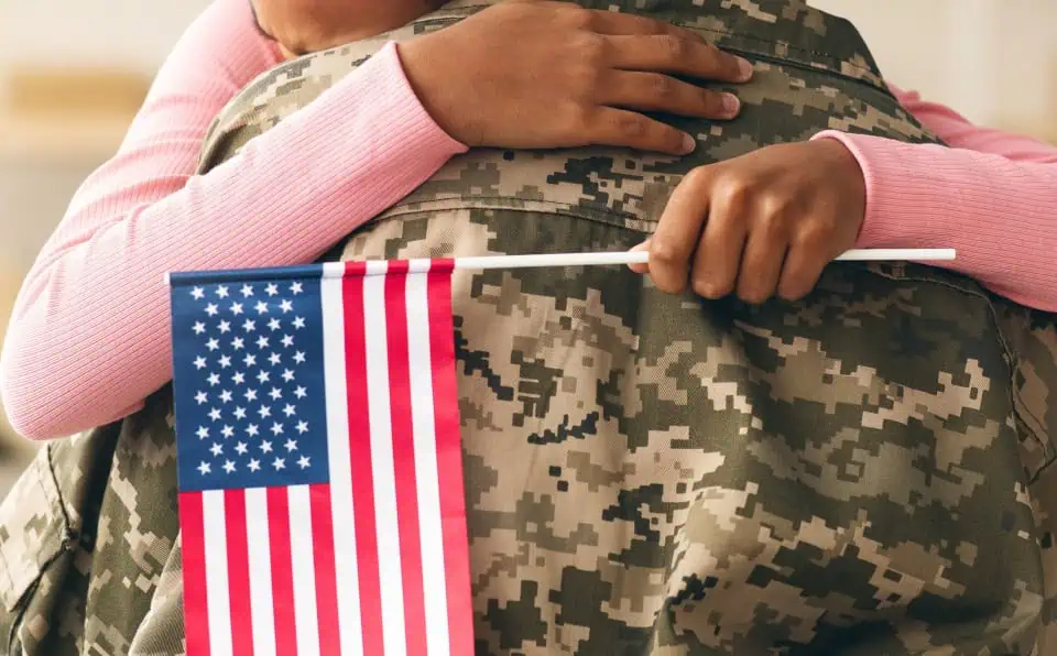 Person in pink sweater hugging a soldier in camouflage uniform holding an American flag.