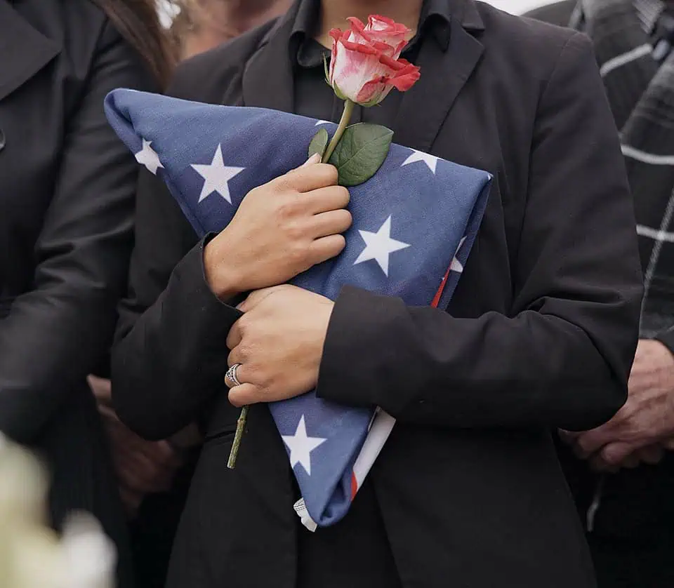 Person holding folded American flag and pink rose at memorial service.