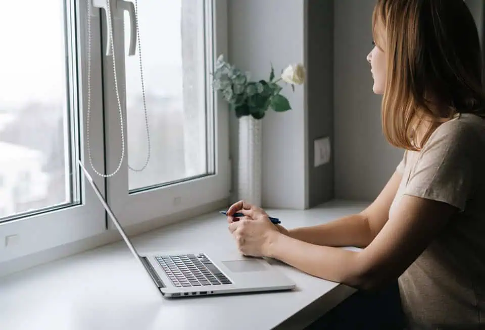 Woman sitting by window with laptop and smartphone in a home setting.