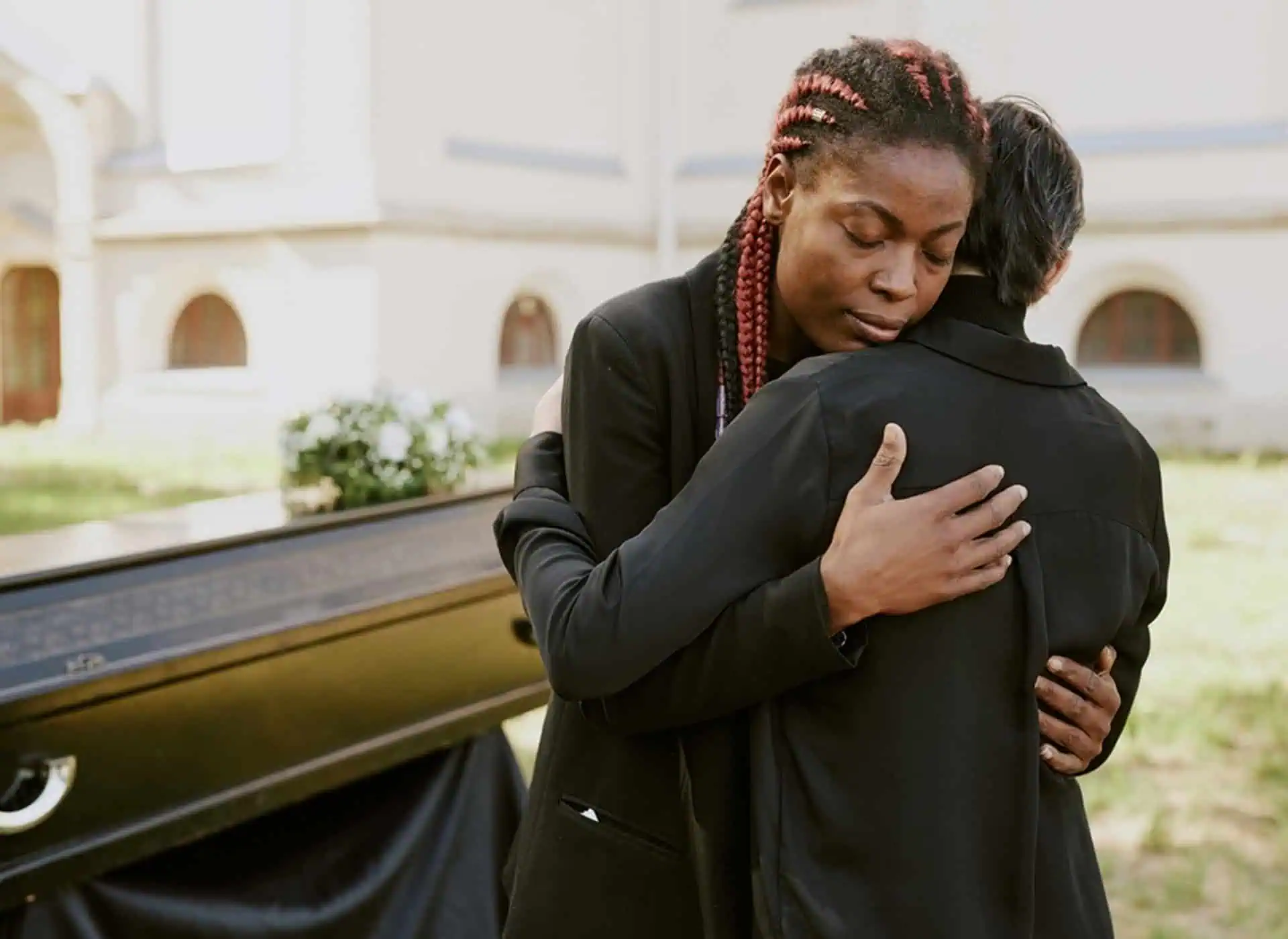 Two women hugging outdoors at a memorial park with a building in the background.